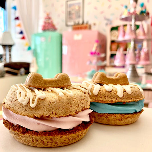 Two decorated dog treats like sponge cake sandwiches with pink and blue cream filling, topped with white drizzle and a heart-shaped biscuit, set on a table in a colorful, blurred kitchen background.