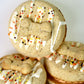 Three round dog treats decorated with white icing, colorful sprinkles, and a small bone-shaped piece in the center, stacked together on a light background.